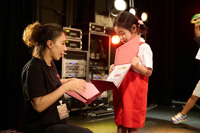 舞台裏で賞状を受け取る赤い制服の女の子とスタッフ